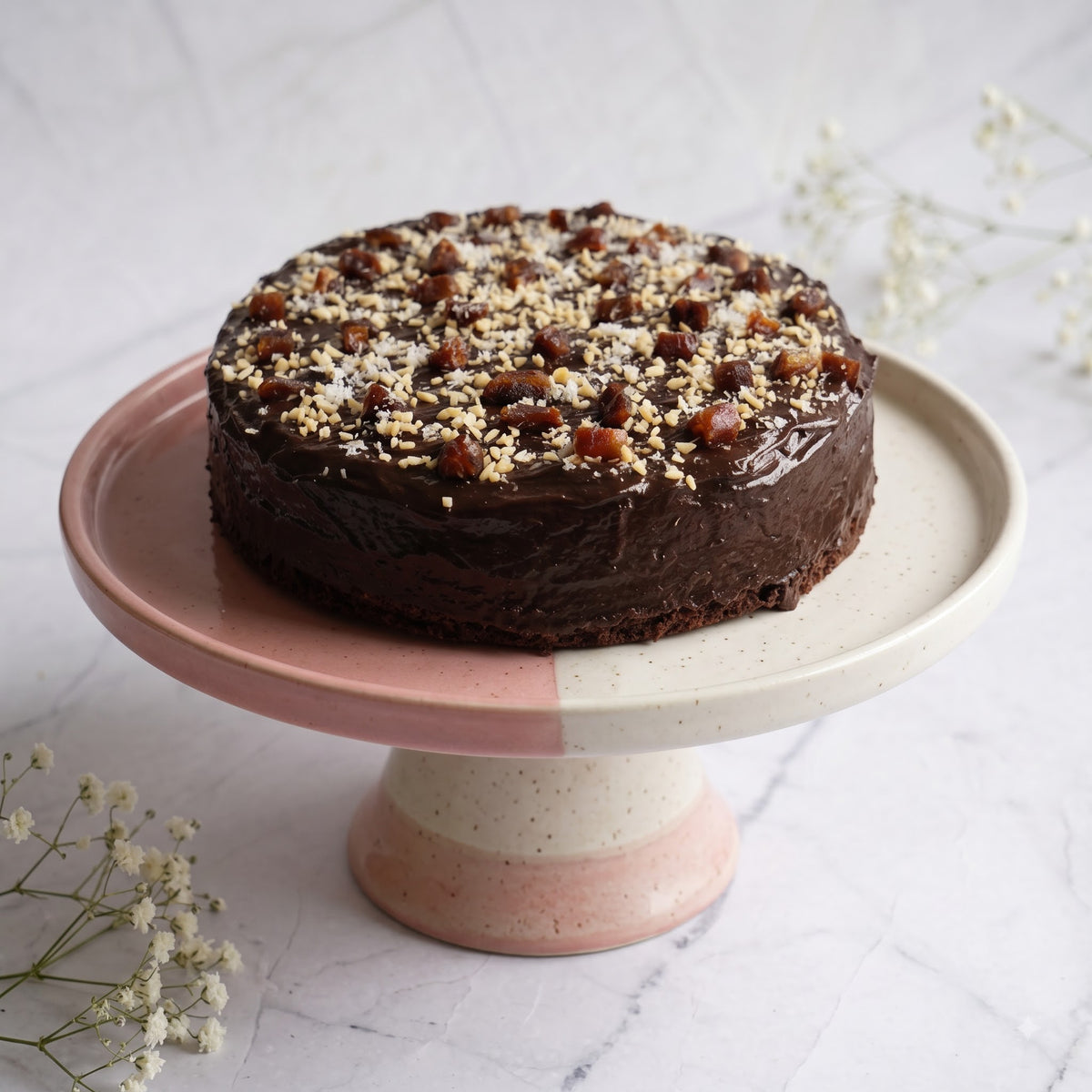 A round chocolate cake with glossy chocolate frosting and chopped nuts on top, displayed on a pink and cream ceramic cake stand against a light marble surface with small white flowers.